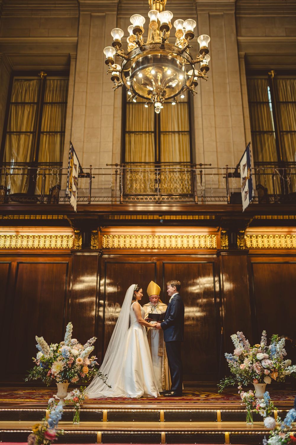 Elegant wedding ceremony in a grand hall with chandeliers and floral arrangements.