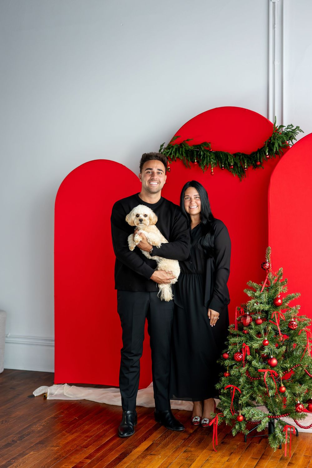 Couple in matching black outfits holding a fluffy white dog in front of red arch backdrop with Christmas tree.