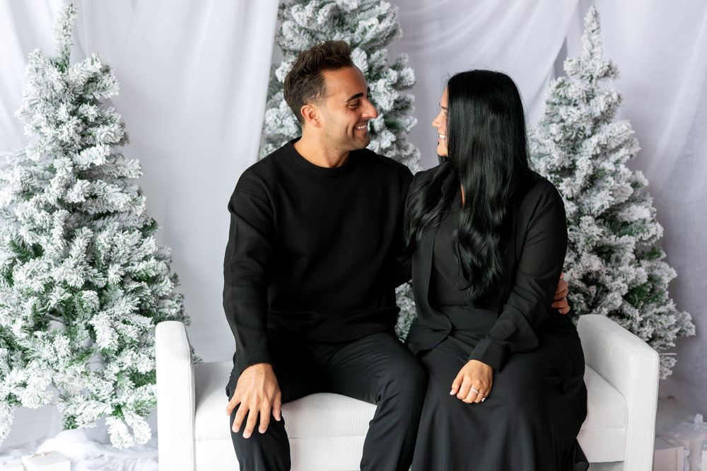 Couple in black outfits gazing at each other lovingly while seated among snow-flocked Christmas trees.