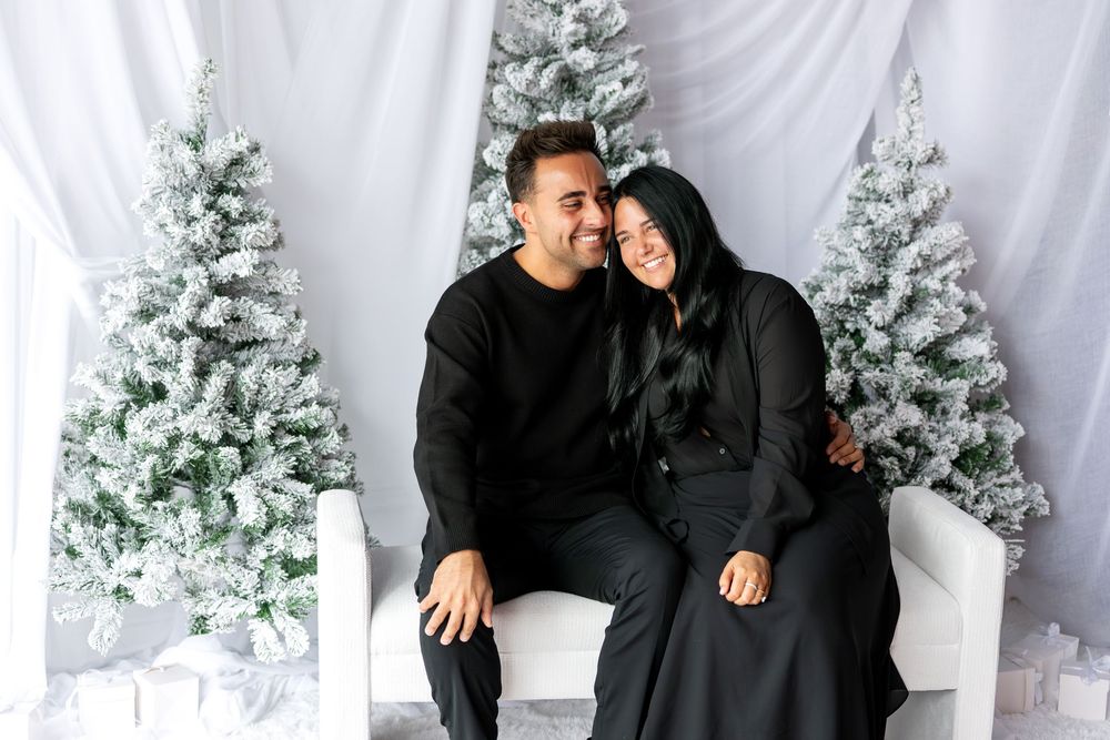 Happy couple in matching black outfits cuddling together on white bench with snowy Christmas trees behind them.
