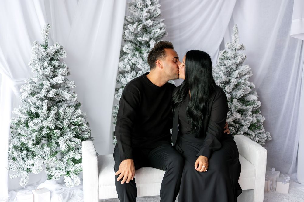 Romantic couple in black outfits sharing a kiss on white bench surrounded by snow-flocked Christmas trees.