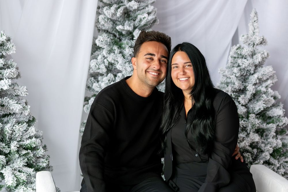 Smiling couple in black outfits posing together on white bench with snow-flocked Christmas trees in background.