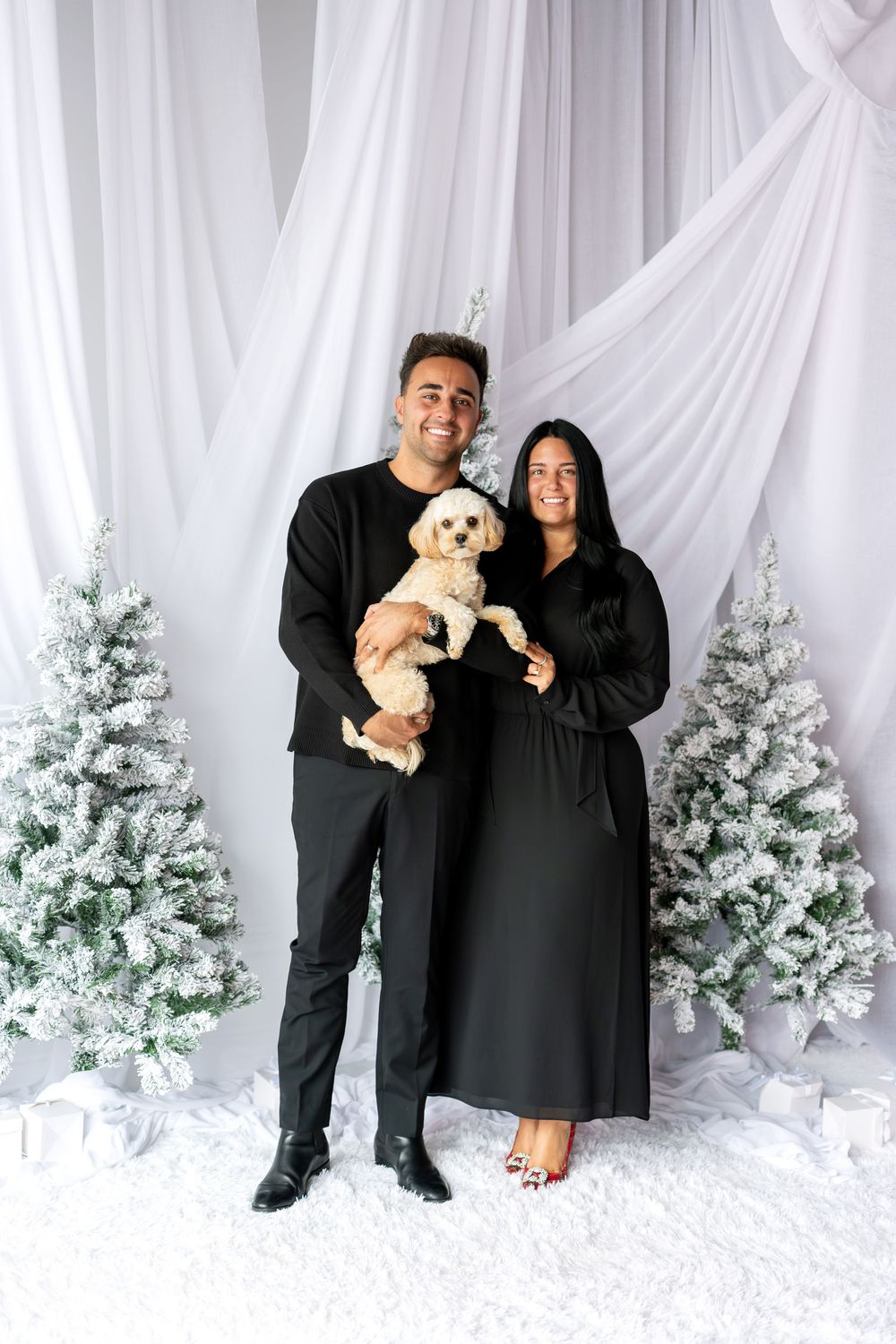 Couple in matching black outfits holding a fluffy cream dog in front of snowy Christmas trees.