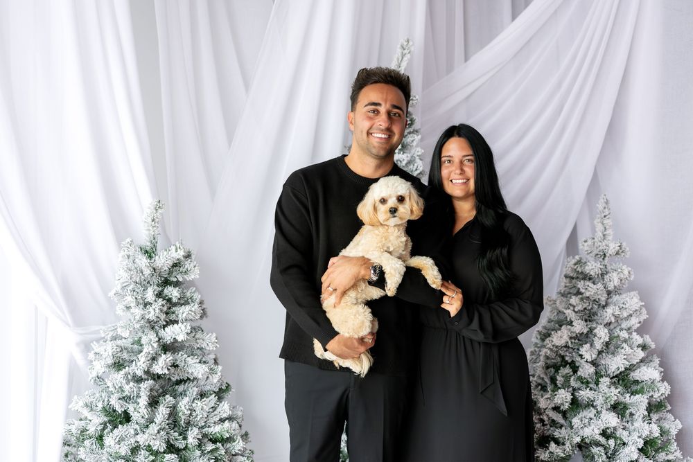Smiling couple dressed in black holding a small cream poodle mix before flocked Christmas trees.
