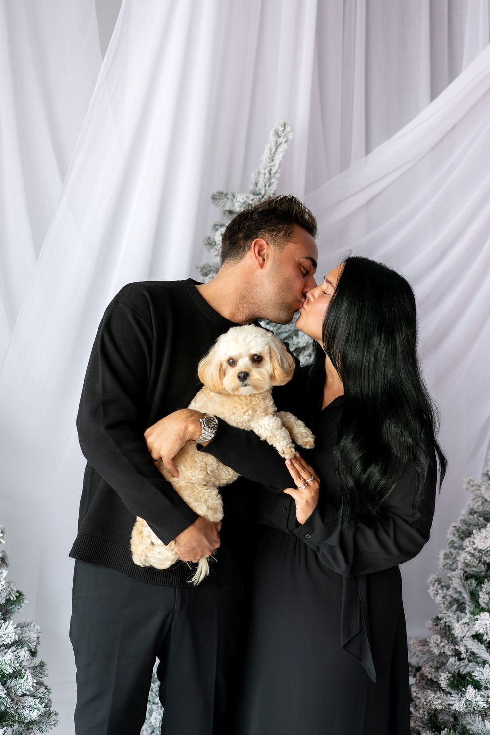 Couple in black outfits kissing while holding a cream poodle mix near snowy Christmas trees.