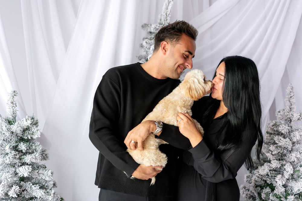 Woman letting small cream poodle lick her face while man smiles near flocked Christmas trees.