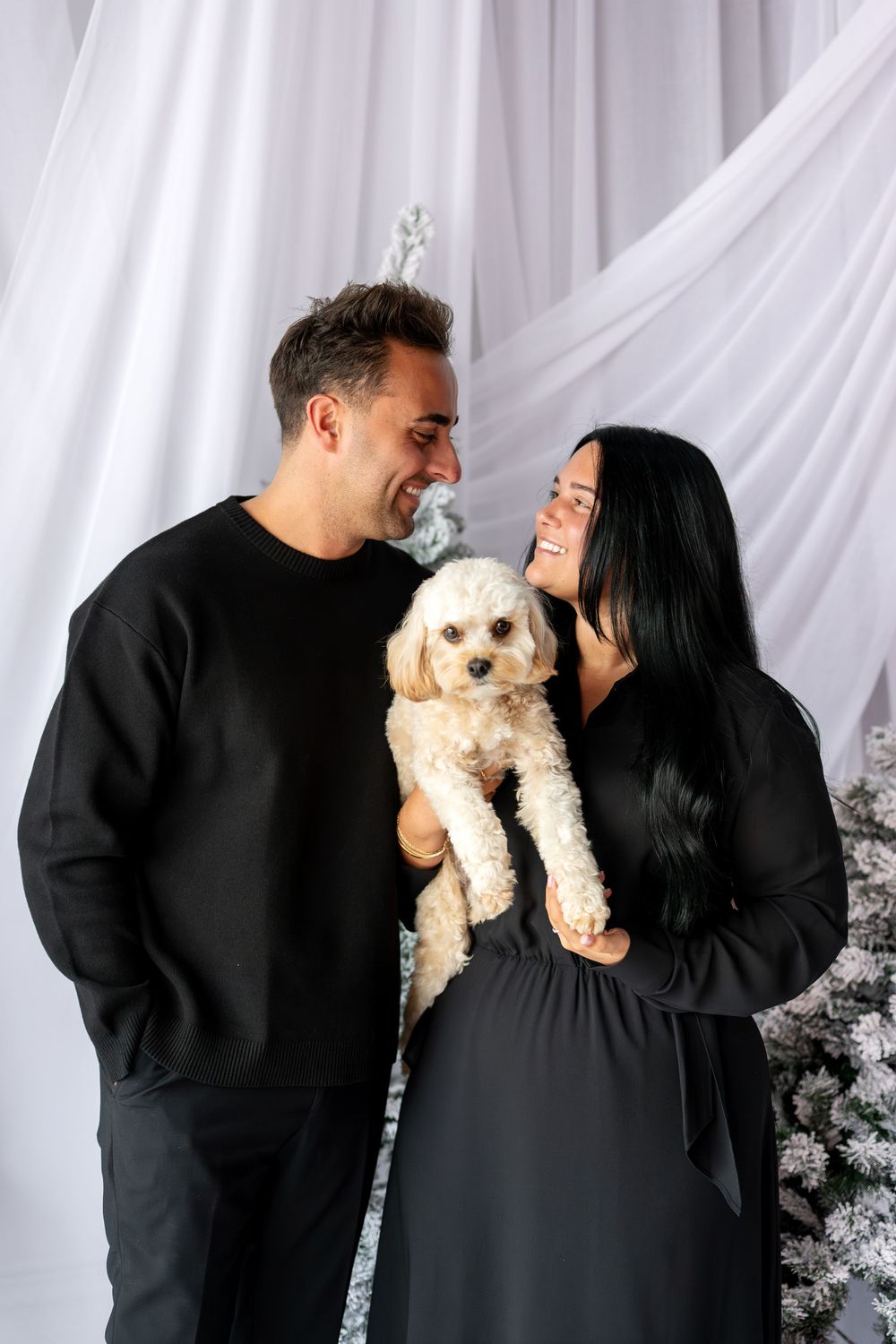 Happy couple in black outfits sharing a smile while holding their cute cream poodle dog.