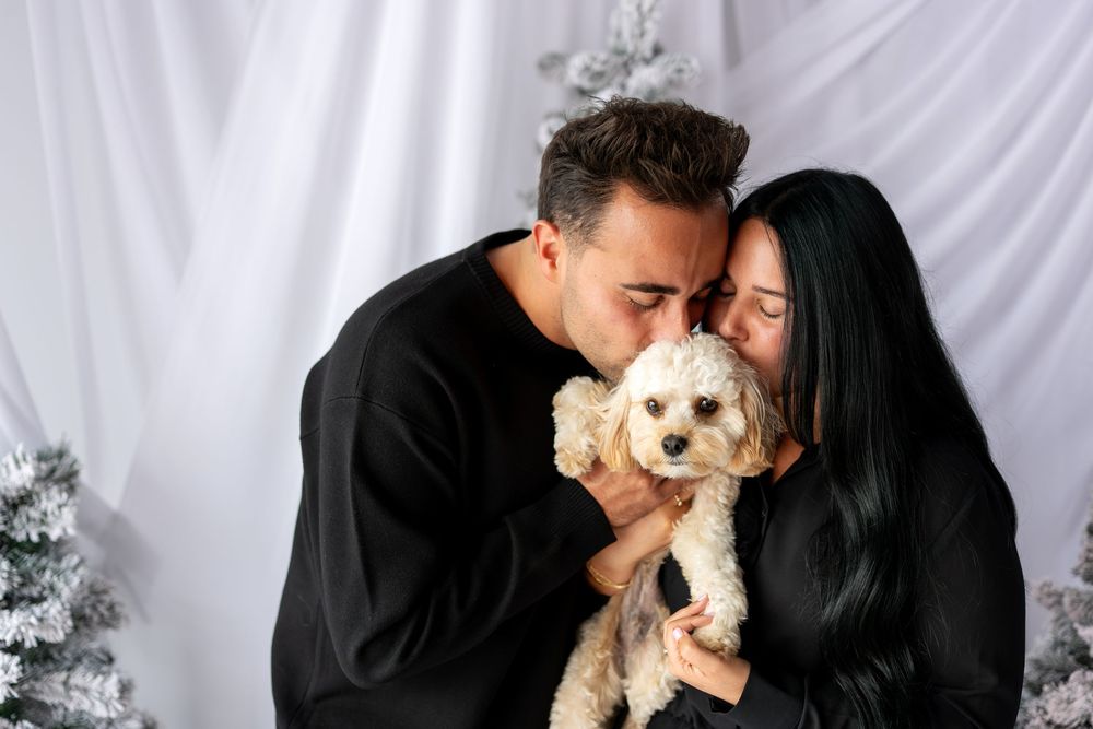 Couple in black outfits kissing their fluffy cream poodle mix near snow-covered Christmas trees.