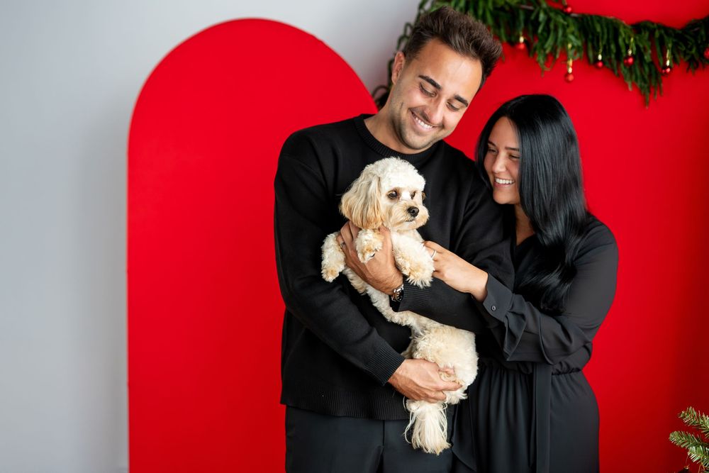 Smiling couple in black clothing laughing together while holding an adorable cream-colored fluffy dog at Christmas.