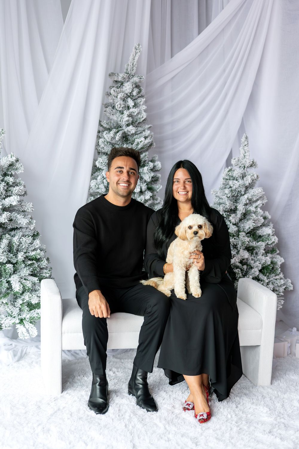 Couple in black outfits sitting with fluffy dog before snow-flocked Christmas trees.