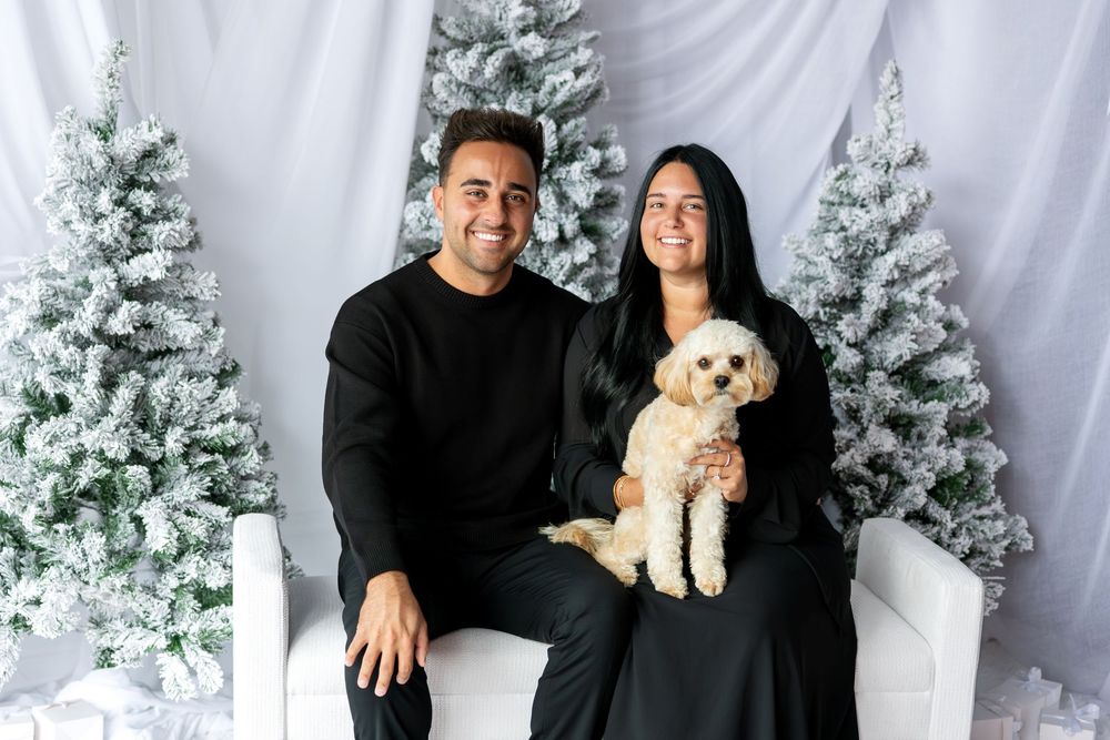 Smiling couple in black outfits holding fluffy dog before snow-flocked Christmas trees.