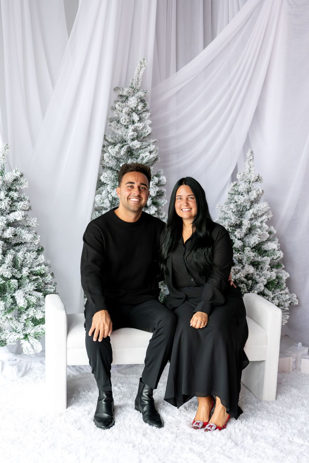 Smiling couple dressed in black sitting together on white bench surrounded by snow-flocked Christmas trees.