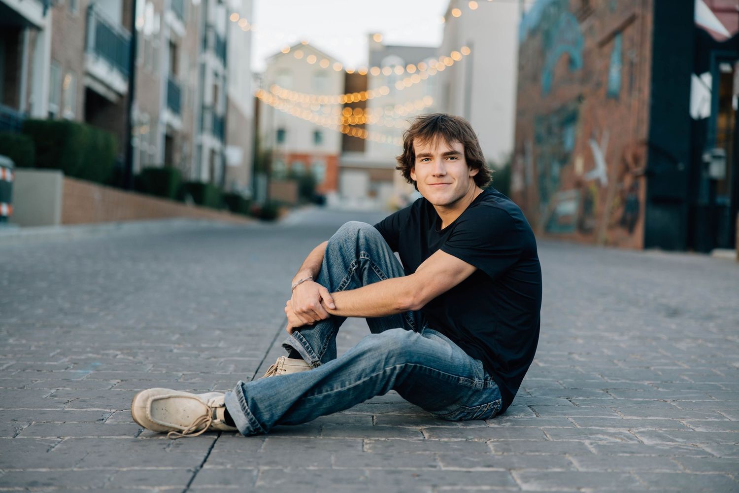 Young man in black t-shirt sitting relaxed on cobblestone street at sunset.