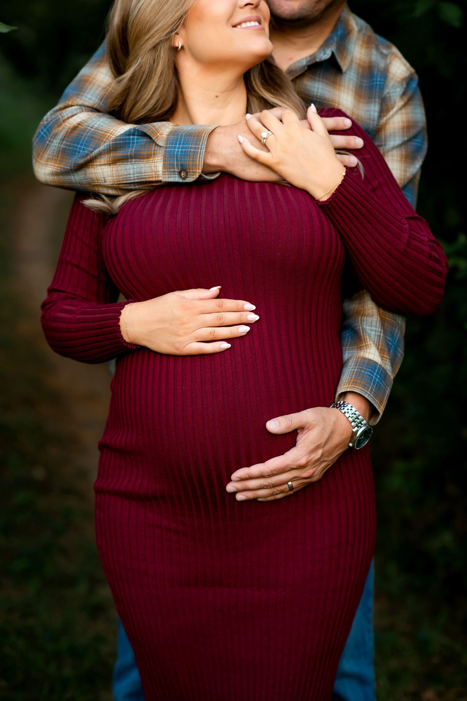 A couple embraces lovingly in a romantic maternity outdoor photoshoot, with the woman wearing a burgundy dress.