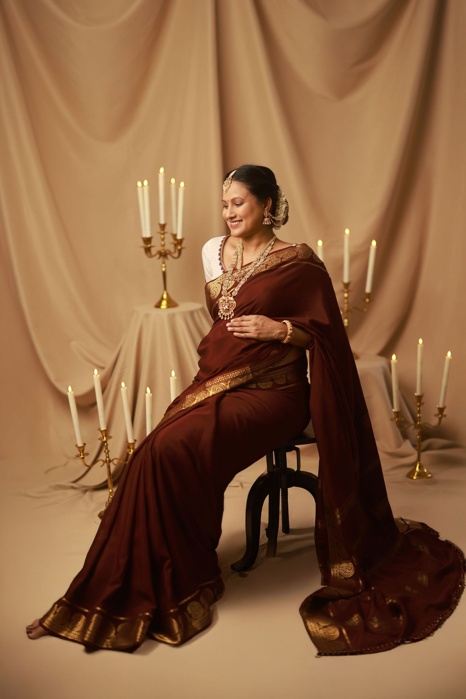 A figure in a flowing brown dress poses gracefully against a beige backdrop with elegant candelabras creating a romantic atmosphere.