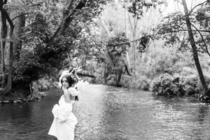 Artistic black and white wedding photo by a tranquil stream with trees framing the scene.