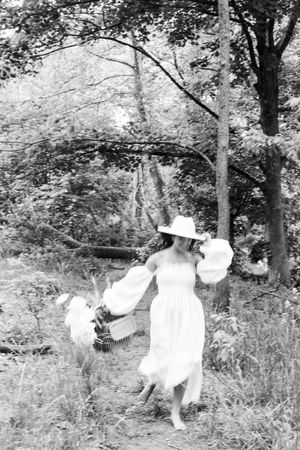 A dreamy black and white photo of someone in a white dress and sun hat dancing in a forest clearing.