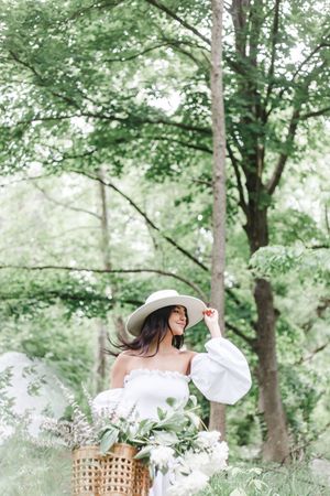Elegant summer photoshoot featuring a wide-brimmed hat and white dress among leafy trees.
