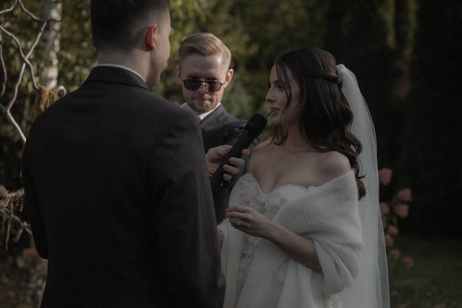 A bride in a strapless white wedding dress and long veil poses outdoors against a natural wooded background.