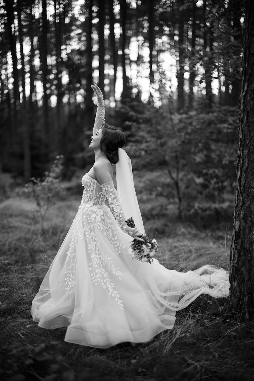 Black and white photo of bride in lace gown reaching upward among forest pine trees.