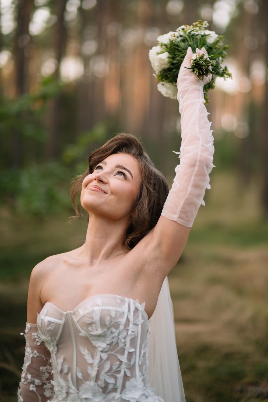 Smiling bride in lace gown raises bouquet of white flowers in sunlit forest.