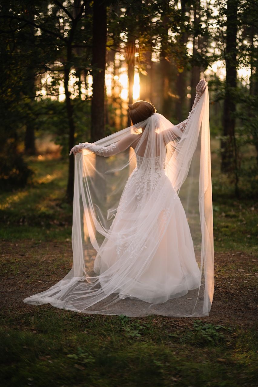 Bride in lace gown spreads flowing veil wide in golden sunset forest setting.