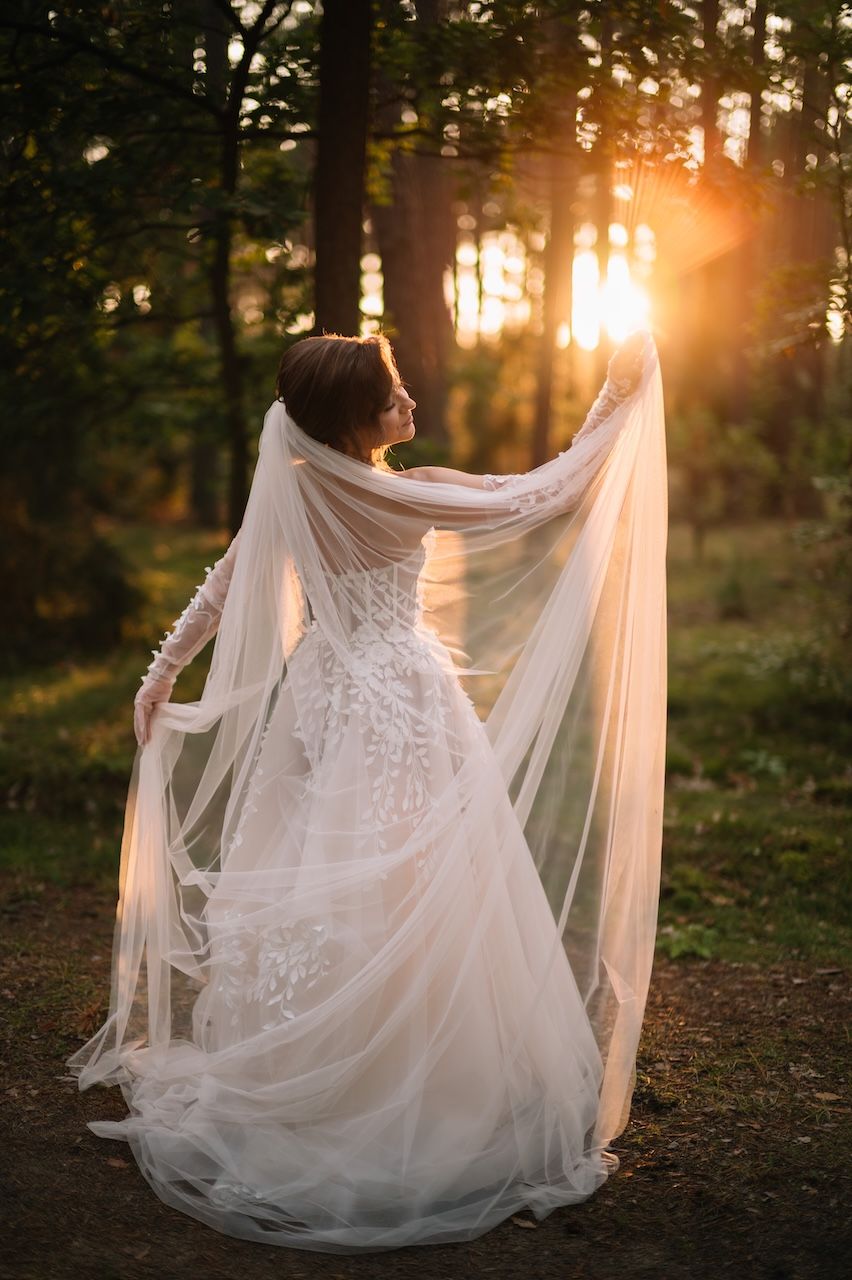 Bride in floral lace gown holds veil aloft with warm sunset light in forest.