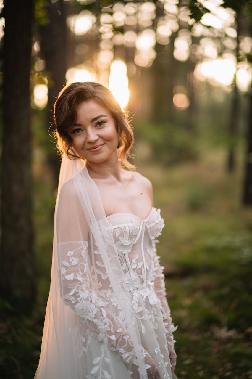 Smiling bride in lace wedding gown poses in golden hour forest light.