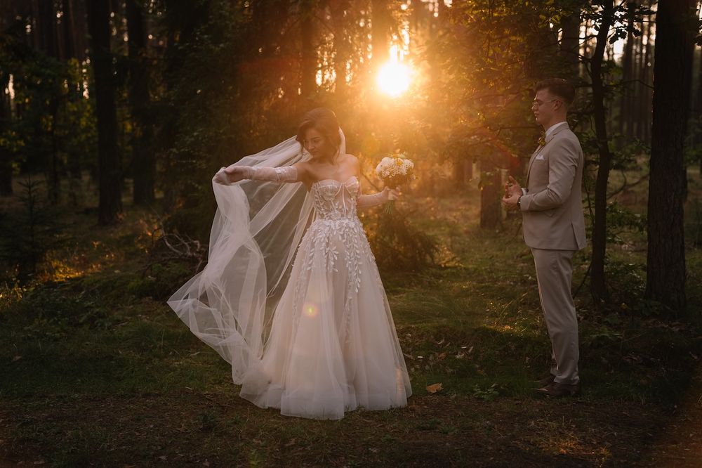 Bride and groom stand in glowing golden sunset light among tall forest trees.