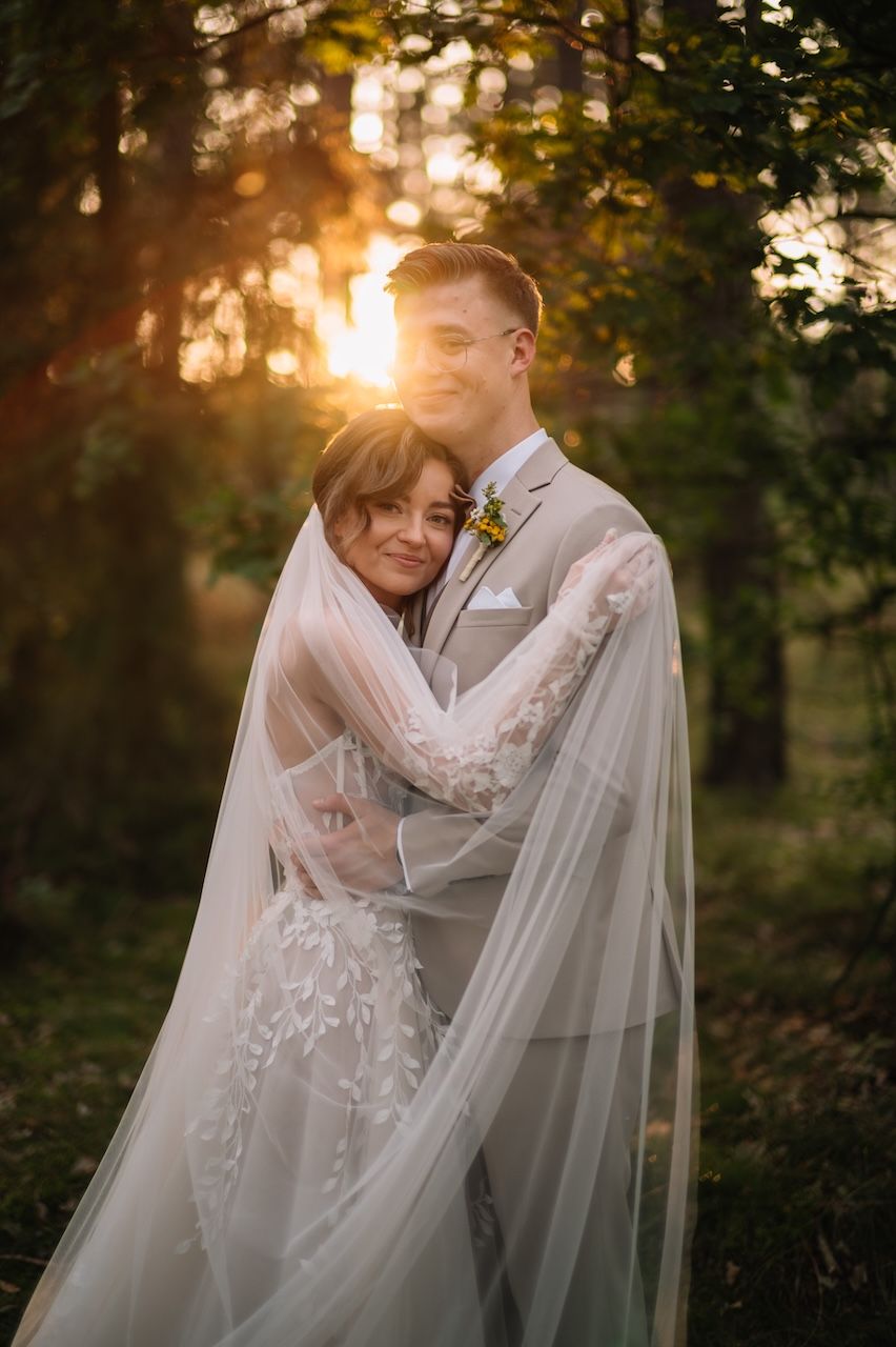 Bride and groom embrace tenderly wrapped in veil with golden forest sunset behind.