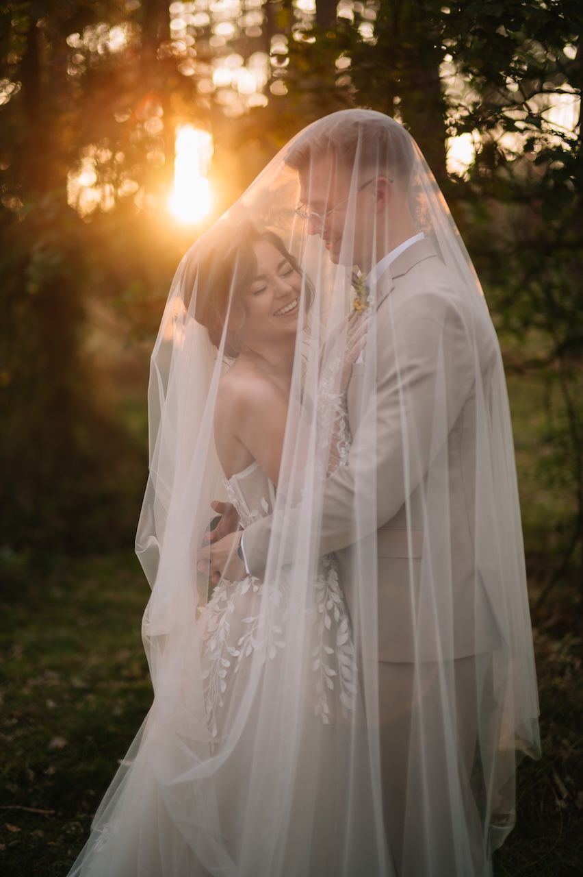 Bride and groom share an intimate moment under a flowing veil with golden sunset light filtering through forest trees.