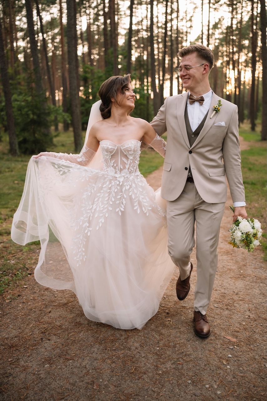Smiling bride and groom walk hand in hand through a pine forest path at golden hour sunset.