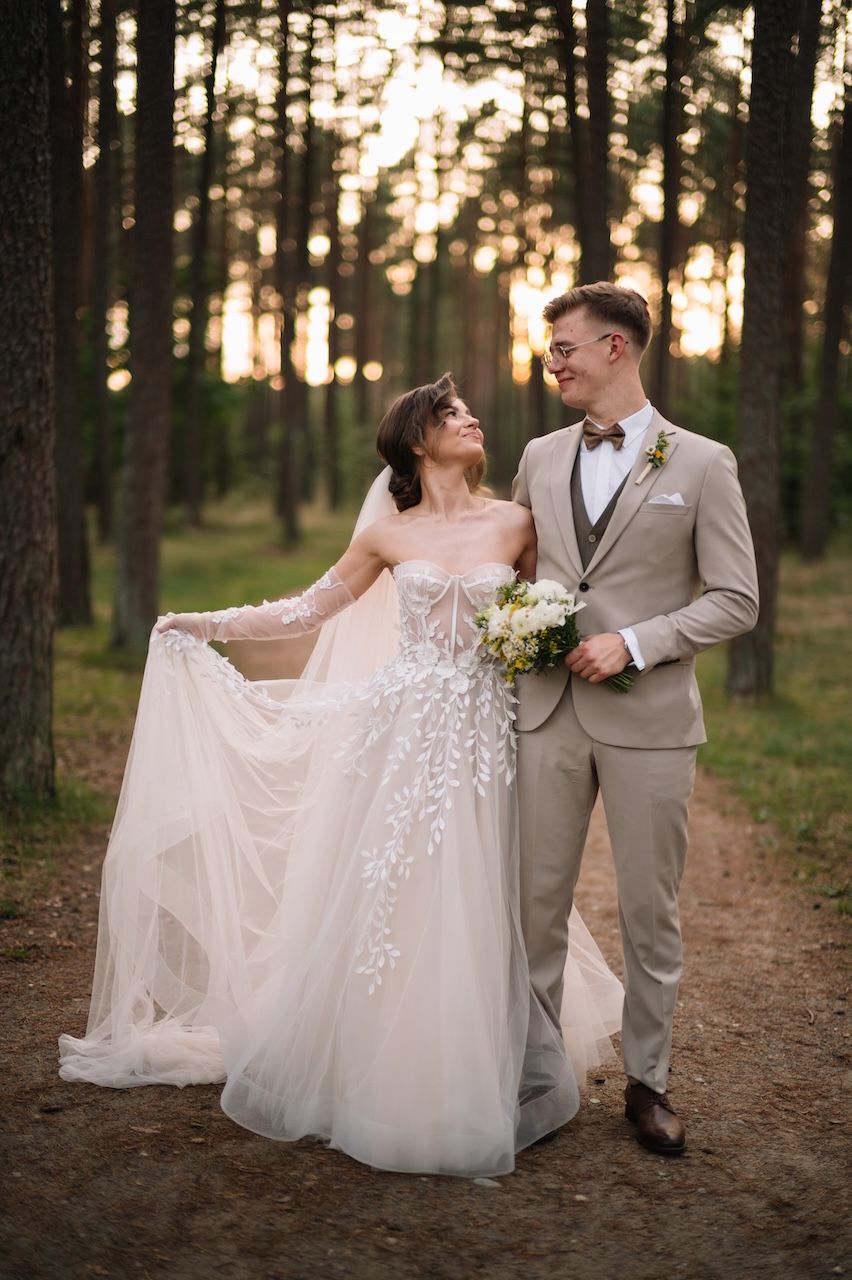 Elegant bride and groom pose together on a forest path at sunset, bride holding her floral lace gown.