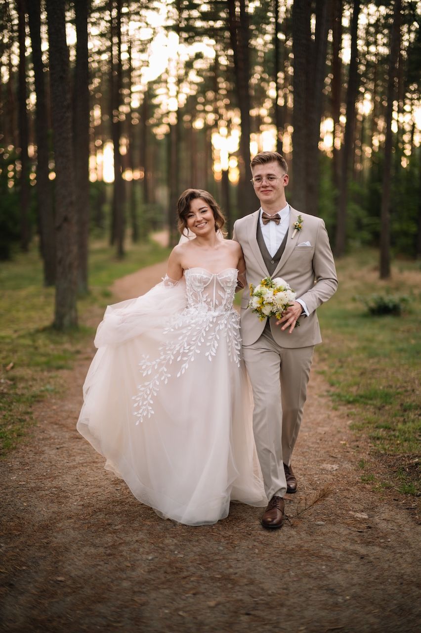 Happy bride and groom stroll along a sunlit pine forest path, groom holding a white wedding bouquet.