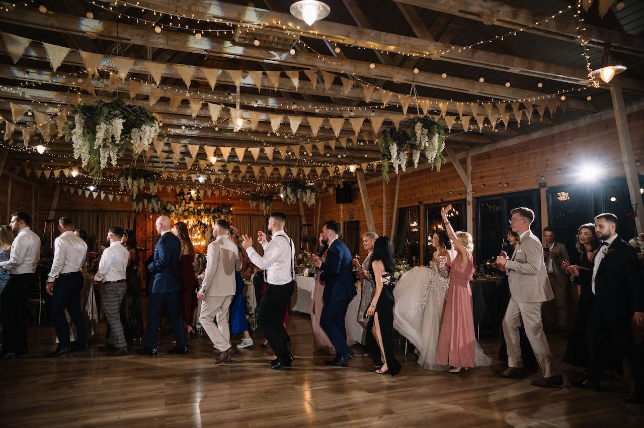 Wedding guests line dance together in a beautifully decorated rustic barn reception venue with string lights.