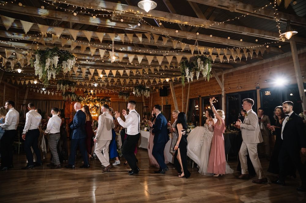 Wedding guests line dance together in a beautifully decorated rustic barn reception venue with string lights.