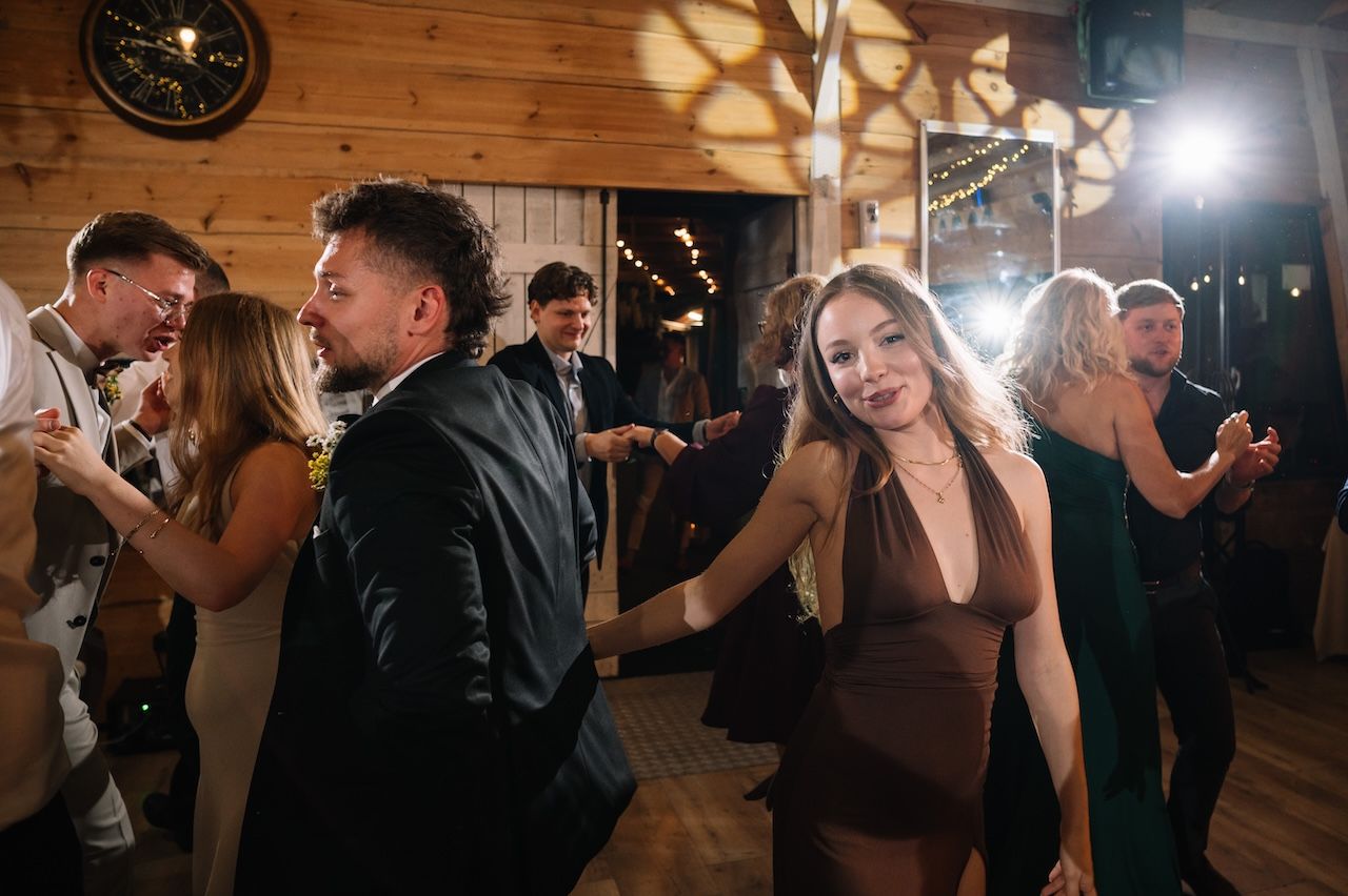Wedding guests dance and celebrate at a rustic barn reception with warm ambient lighting.