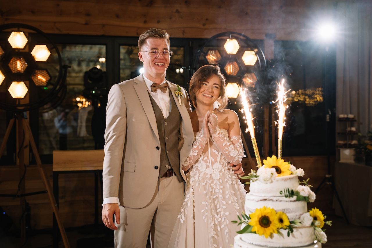 Bride and groom smile beside a sunflower wedding cake with sparkling sparklers at their reception.