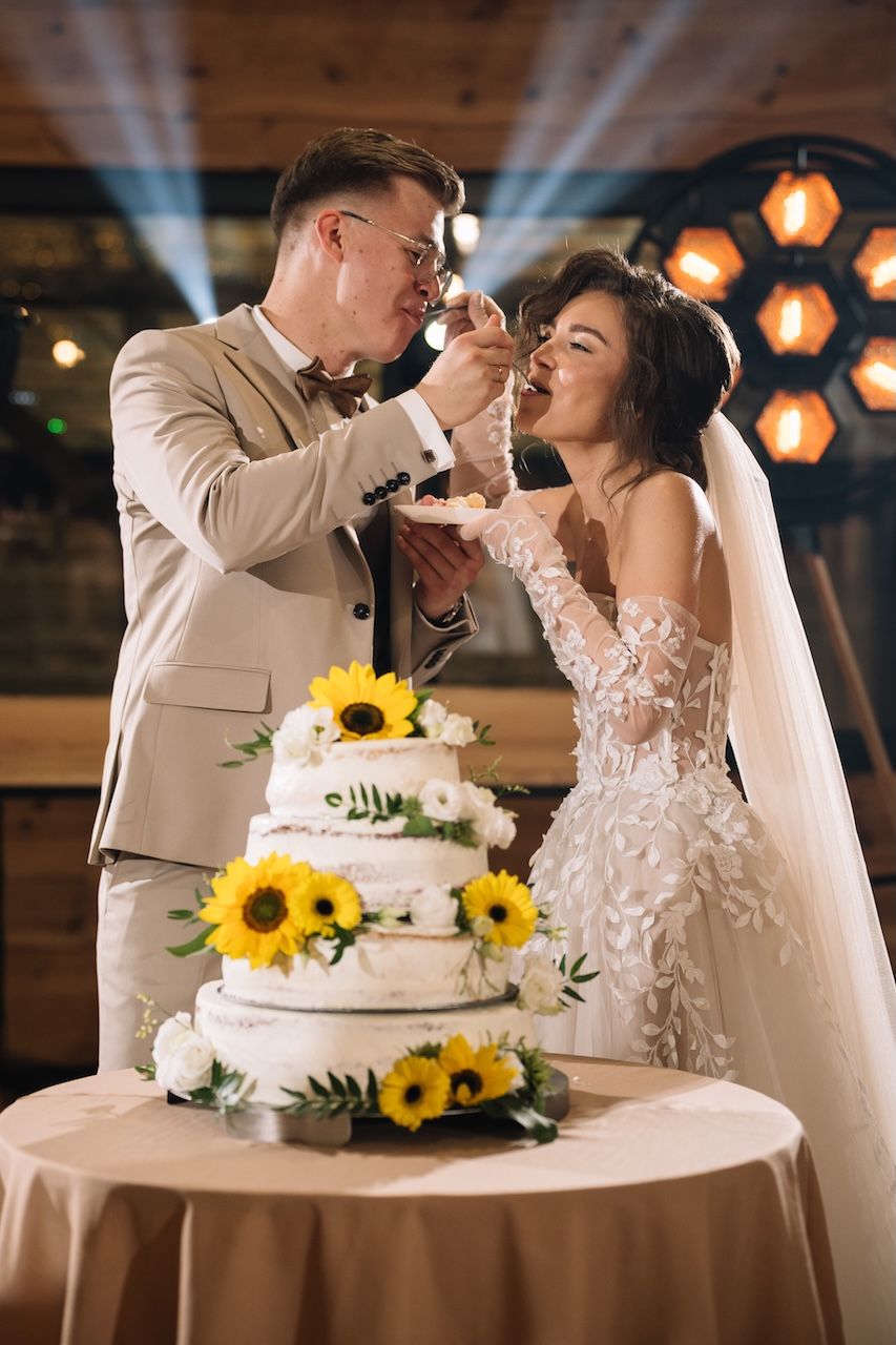 Bride and groom feed each other wedding cake at a reception beside a sunflower-decorated tiered cake.