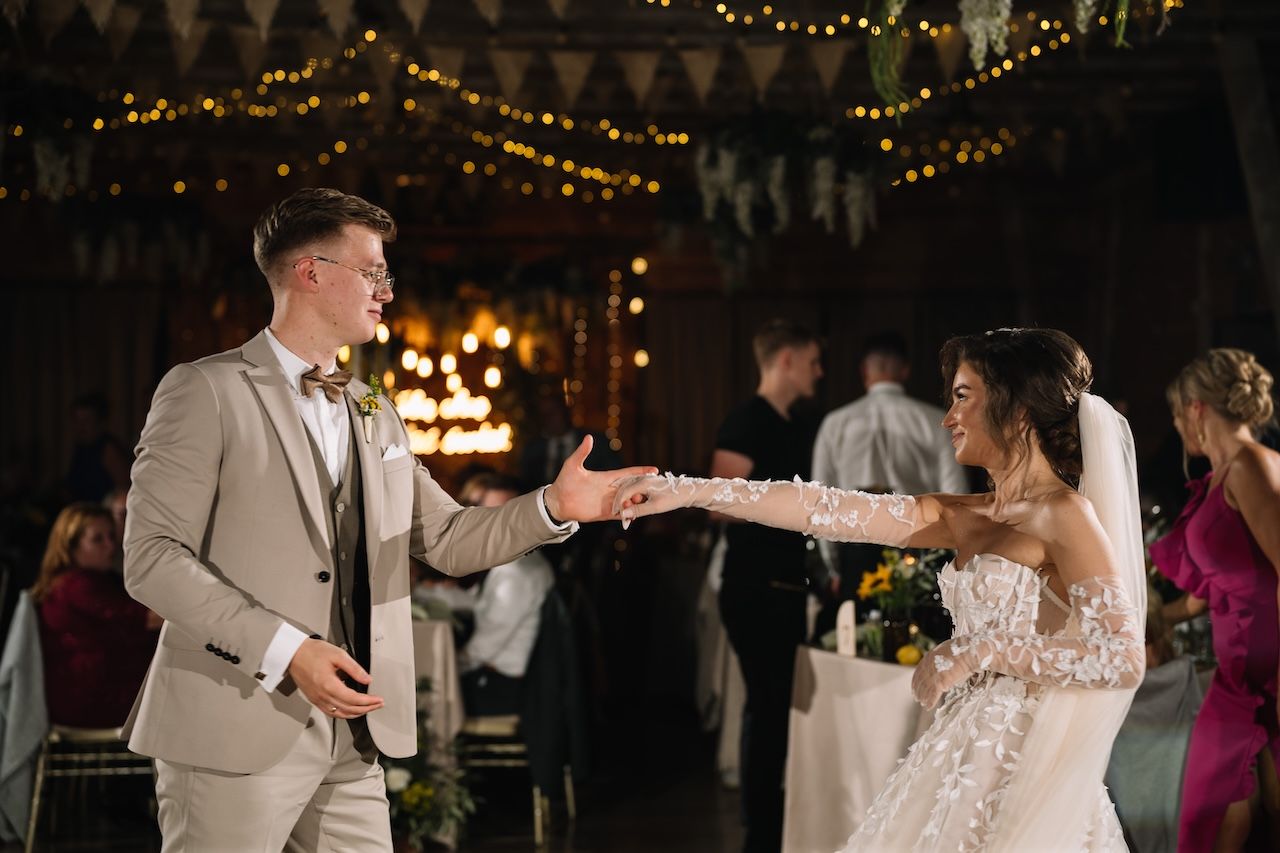Bride and groom share their first dance, holding hands under warm fairy lights in a rustic wedding venue.