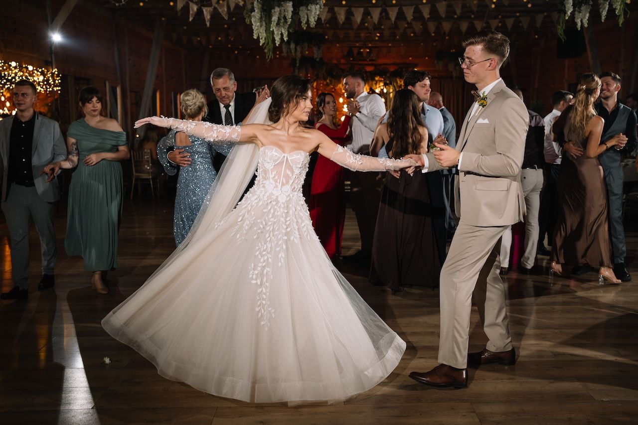 Bride in a floral lace gown twirls elegantly with groom in beige suit during wedding reception dance.