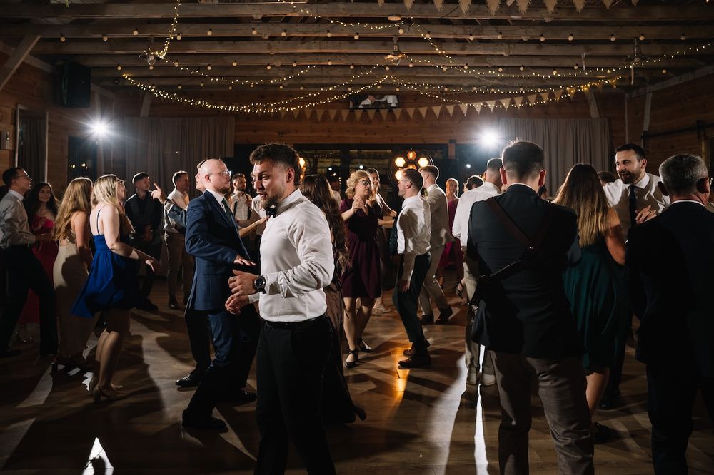 Wedding guests fill the dance floor, celebrating joyfully under string lights and bunting in a barn venue.
