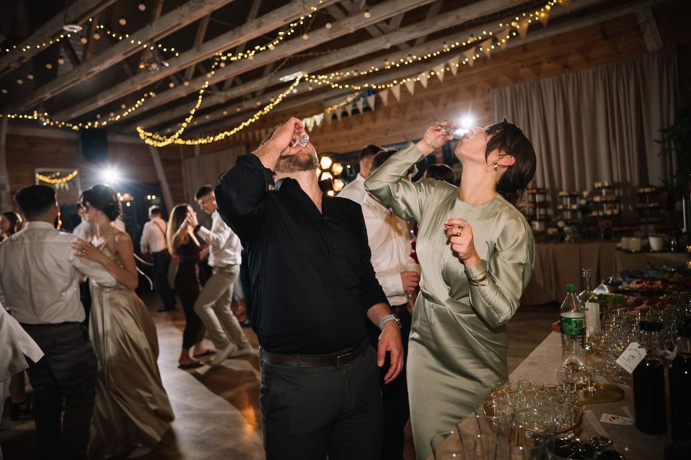 Wedding guests dance and take photos under fairy lights at a lively rustic barn reception celebration.