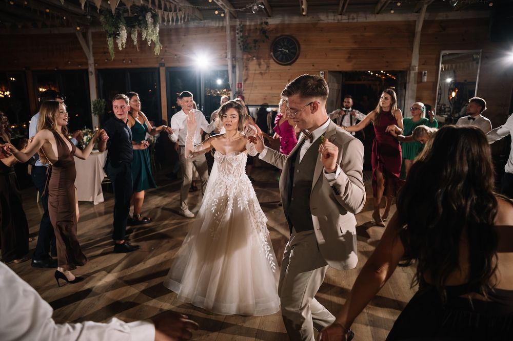 Bride in lace gown and groom dance joyfully surrounded by celebrating guests at rustic barn wedding.
