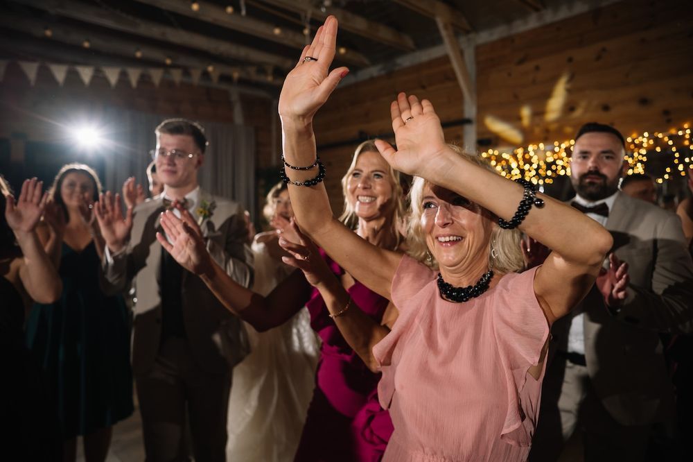 Enthusiastic wedding guest dances with arms raised while guests clap and celebrate at barn reception.
