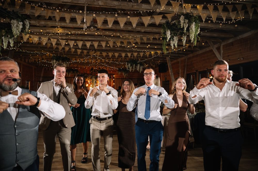 Groomsmen and male wedding guests perform a coordinated fun dance move at a rustic barn reception.