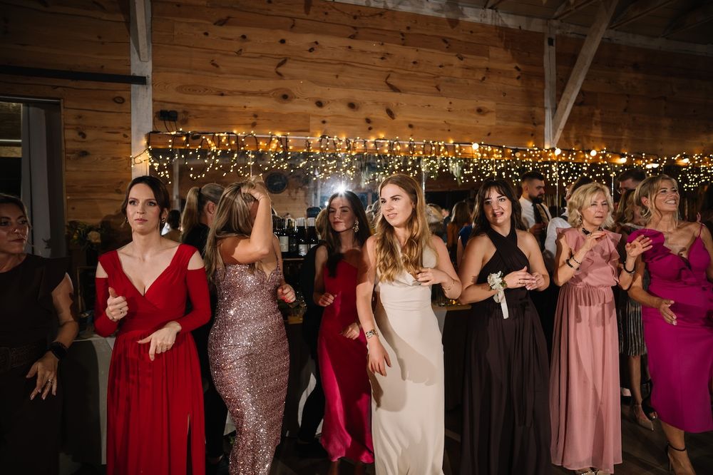 Elegantly dressed female wedding guests dance together in colorful gowns at a festive barn reception.