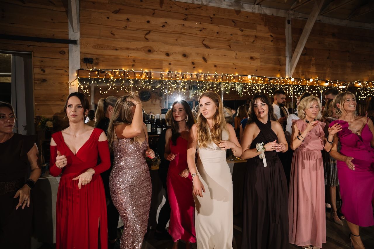 Elegantly dressed female wedding guests dance together in colorful gowns at a festive barn reception.