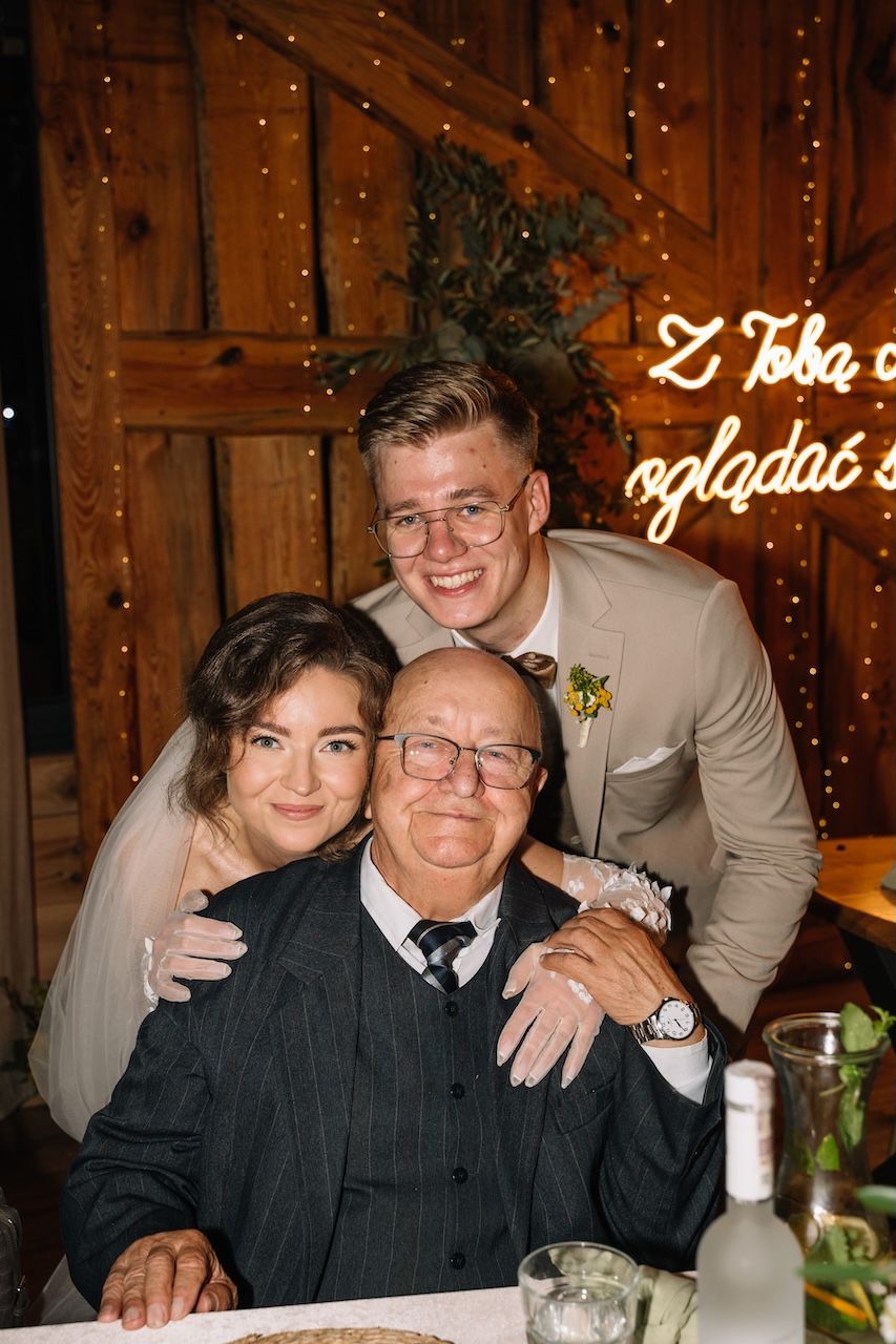 Newlyweds pose warmly with elderly grandfather at wedding table beneath a glowing neon sign in barn.