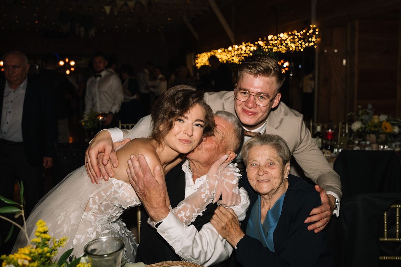 Bride and groom pose with elderly grandparents in joyful wedding reception family photo.